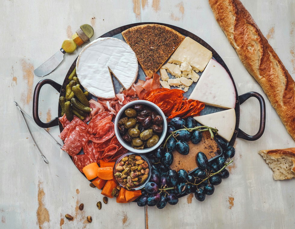 The full charcuterie board from above — brie, aged cheddar, folded salami, olives, grapes and a torn baguette.
