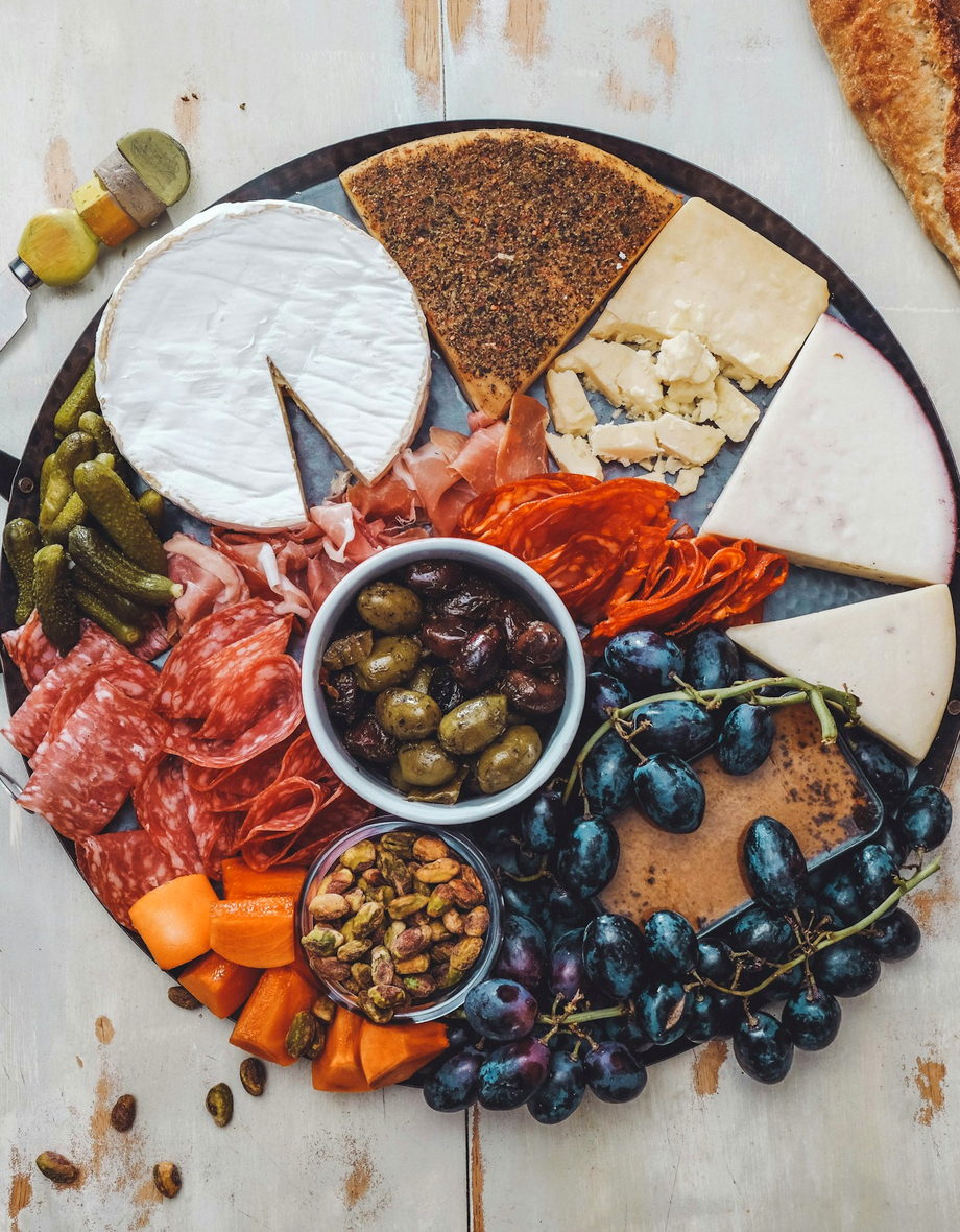 An overhead grazing board crowded with aged cheeses, folded prosciutto, olives, grapes and torn baguette on a pale linen surface.