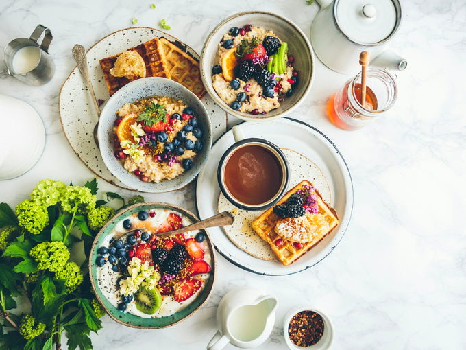 An overhead brunch spread of bowls, fruit, pastries and coffee on a bright table.